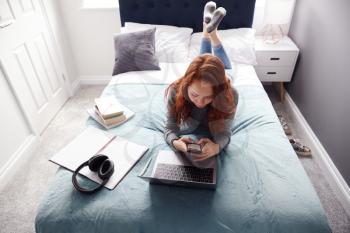 Looking Down On Female College Student Lying On Bed Working On Laptop And Using Mobile Phone