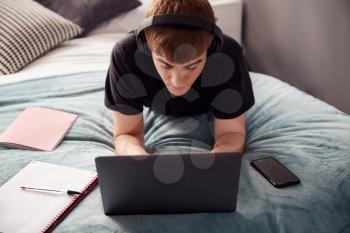 Male College Student Wearing Headphones Lies On Bed In Shared House Working On Laptop