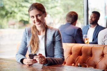 Portrait Of Businesswoman Sitting In Bar Checking Mobile Phones Whilst Colleagues Meet For Drink