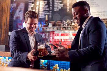 Two Businessmen Checking Mobile Phones Whilst Meeting For Drink In Bar