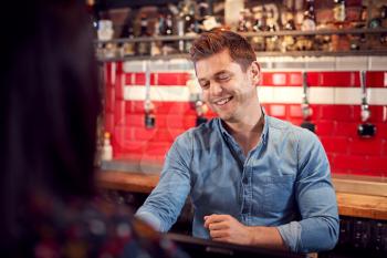 Male Bar Tender Standing Behind Counter Serving Drinks To Customer