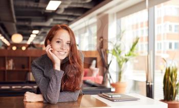 Portrait Of Casually Dressed Young Businesswoman Working At Desk In Modern Open Plan Workplace