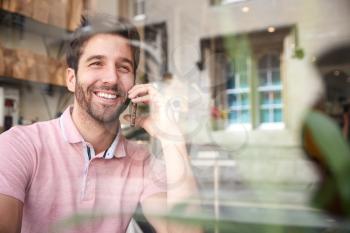 Young Man In Coffee Shop Talking On Mobile Phone Viewed Through Window