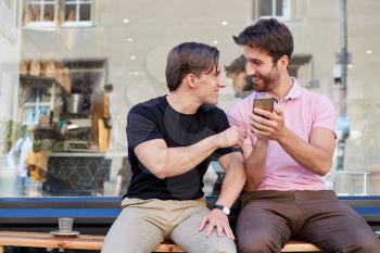 Male Gay Couple Sitting Outside Coffee Shop Looking At Social Media On Mobile Phone