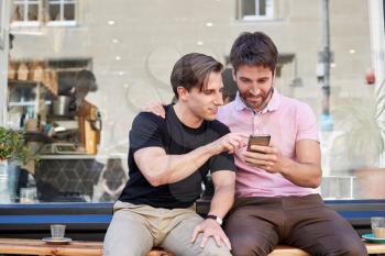 Male Gay Couple Sitting Outside Coffee Shop Looking At Social Media On Mobile Phone