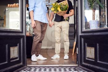 Close Up Of Loving Male Gay Couple Holding Hands Coming Out Of Florists Holding Bunch Of Flowers
