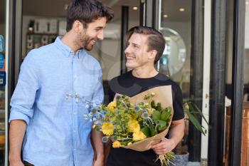Loving Male Gay Couple Holding Hands Coming Out Of Florists Holding Bunch Of Flowers
