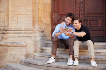 Male Gay Couple On Vacation Sitting Outdoors On Steps Of Building Looking At Mobile Phone