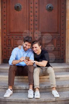Male Gay Couple On Vacation Sitting Outdoors On Steps Of Building Looking At Mobile Phone