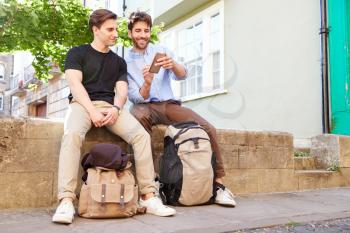 Male Gay Couple On Vacation Sitting On Wall With Backpacks Looking At Mobile Phone
