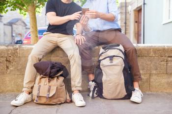 Close Up Of Male Gay Couple On Vacation Sitting On Wall With Backpacks Looking At Mobile Phone