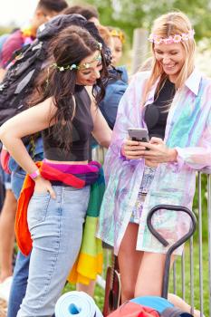 Female Friends Look At Mobile Phone As They Wait Behind Barrier At Entrance To Music Festival Site