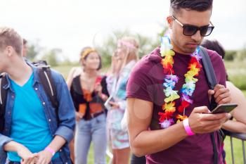 Young Man Looking At Mobile Phone As He Waits Behind Barrier At Entrance To Music Festival Site