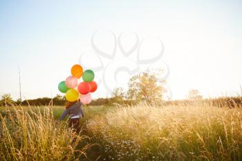 Rear View Of Two Female Friends Camping Running Through Field With Balloons Against Flaring Sun