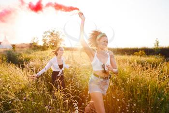 Two Female Friends Camping At Music Festival Running Through Field With Smoke Flare