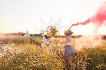 Two Female Friends Camping At Music Festival Running Through Field With Smoke Flares
