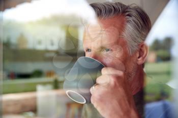 Senior Man Standing And Looking Out Of Kitchen Door Drinking Coffee Viewed Through Window