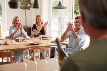 Senior Man Opens Bottle Of Champagne As Family With Adult Offspring Eat Meal Around Table At Home
