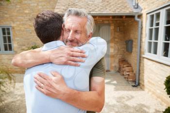 Senior Father Greets And Hugs Adult Son Outside Front Door Of House As He Visits