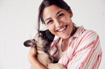 Studio Portrait Of Smiling Young Woman Holding Affectionate Pet French Bulldog Puppy