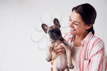 Studio Shot Of Smiling Young Woman Holding Affectionate Pet French Bulldog Puppy