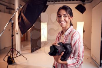 Portrait Of Smiling Female Photographer Standing In Studio With Camera And Lighting Equipment