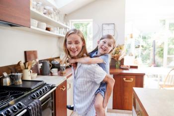 Mother Giving Smiling Daughter Piggyback Ride In Kitchen At Home
