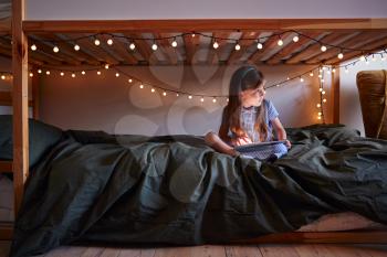 Young Girl Sitting On Bed At Home Decorated With Fairy Lights Using Digital Tablet