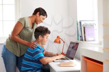 Mother Helping Son In Bedroom To Do Homework Using Laptop On Desk