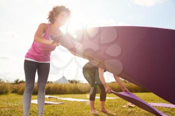 Two Mature Female Friends Laying Out Exercise Mats On Grass At Outdoor Yoga Retreat