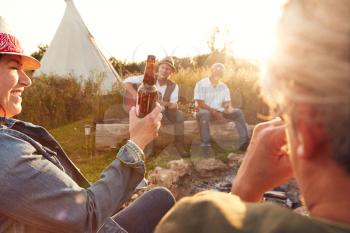 Group Of Mature Friends Sitting Around Fire As They Drink And Sing Songs At Outdoor Campsite