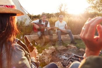 Group Of Mature Friends Sitting Around Fire As They Drink And Sing Songs At Outdoor Campsite