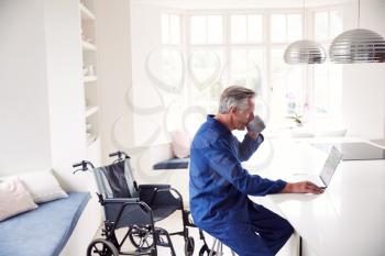 Mature Disabled Man In Wheelchair At Home Using Laptop On Kitchen Counter