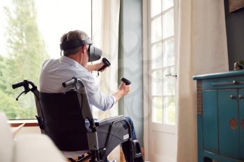 Mature Disabled Man In Wheelchair At Home Wearing Virtual Reality Headset Holding Gaming Controllers