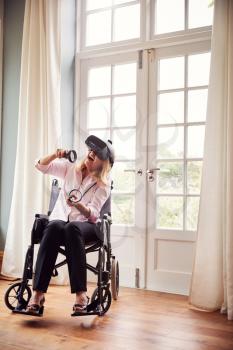Mature Disabled Woman In Wheelchair At Home Wearing Virtual Reality Headset With Gaming Controllers