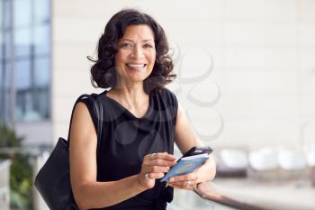 Portrait Of Mature Businesswoman With Passport In Airport Departure Lounge Using Mobile Phone