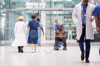 Female Nurse Wearing Scrubs Wheeling Patient In Wheelchair Through Lobby Of Modern Hospital Building