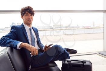 Portrait Of Businessman Sitting In Airport Departure Lounge Using Mobile Phone