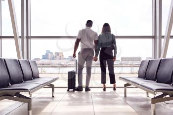 Rear View Of Business Couple With Luggage Standing By Window In Airport Departure Lounge
