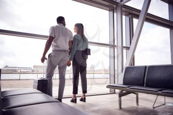 Rear View Of Business Couple With Luggage Standing By Window In Airport Departure Lounge