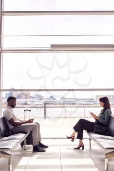 Businessman And Businesswoman Sitting In Airport Departure Lounge Using Mobile Phone