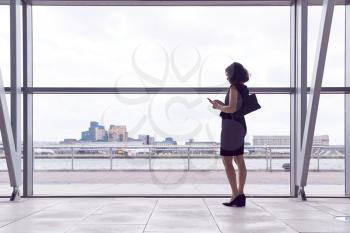 Rear View Of Businesswoman Using Mobile Phone Standing By Window In Airport Departure Lounge