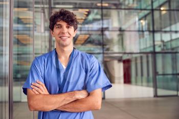 Portrait Of Young Male Doctor Wearing Scrubs Standing In Modern Hospital Building