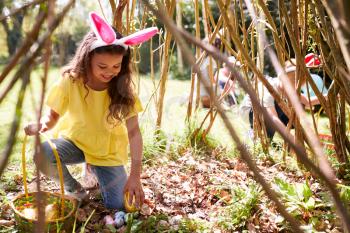 Group Of Children Wearing Bunny Ears Finding Easter Eggs Hidden In Garden