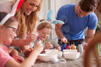 Parents With Children Wearing Bunny Ears Sitting At Table Decorating Eggs For Easter At Home