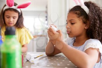 Two Girls Wearing Bunny Ears Sitting At Table Decorating Eggs For Easter At Home