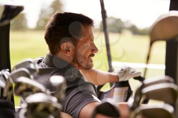 Rear View Of Mature Man Playing Golf Driving Buggy Along Course Viewed Through Golf Clubs