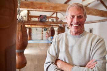 Portrait Of Senior Male Boxing Coach In Gym Standing By Old Fashioned Leather Punching Bags