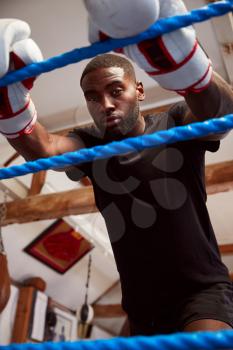 Portrait Of Male Boxer In Gym Wearing Boxing Gloves Leaning On Ropes Of Boxing Ring