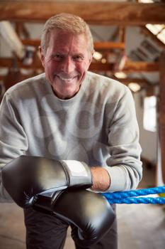 Smiling Senior Male Boxer In Gym Wearing Boxing Gloves Leaning On Ropes Of Boxing Ring